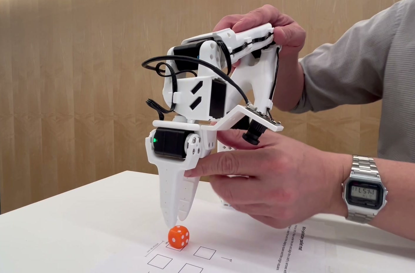 Two hands guiding the SO-ARM101 by hand with torque disabled, lowering the gripper's fingertips toward an orange die sitting on a square of the printed pallet mat.
